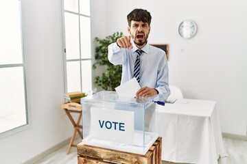 Sticker - Hispanic man with beard voting putting envelop in ballot box pointing displeased and frustrated to the camera, angry and furious with you