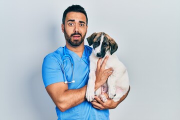 Wall Mural - Handsome hispanic veterinary man with beard checking dog health in shock face, looking skeptical and sarcastic, surprised with open mouth