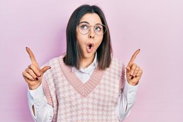 Young brunette woman wearing elegant sweater and glasses amazed and surprised looking up and pointing with fingers and raised arms.