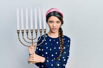 Poster - Young brunette girl holding menorah hanukkah jewish candle clueless and confused expression. doubt concept.