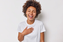 Portrait Of Happy Sincere Woman With Curly Hair Keeps Hand On Chest Expresses Positive Feelings Hears Heartwaring Words Dressed In Casual T Shirt Isolaed Over White Background. Happiness Concept