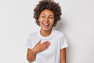 Wall Mural - Portrait of happy sincere woman with curly hair keeps hand on chest expresses positive feelings hears heartwaring words dressed in casual t shirt isolaed over white background. Happiness concept