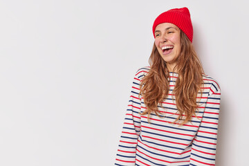 Wall Mural - Beautiful woman with long hair smiles sincerely feels very happy wears casual striped jumper and red hat being in good mood poses against white background blank copy space for your advertisement