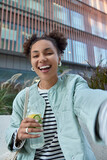 Fototapeta  - Happy optimistic girl with two hair buns dressed in jacket enjoys free time and walking in city holds bottle of detox drink makes selfie poses against modern building has fun during daytime.