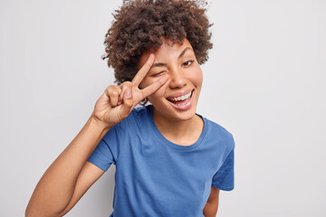 Wall Mural - Glad beautiful woman shows peace sign over eye winks and smiles at camera wears casual blue t shirt isolated over white background demonstrates victory gesture enjoys life. Body language concept