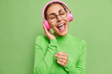 Wall Mural - Carefree positive woman enjoys music in headphones sings favorite song keeps hand as if microphone keeps eyes closed wears spectacles and casual jumper isolated over vivid green studio wall.