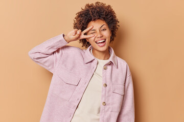 Wall Mural - Positive curly woman winks eye makes peace sign smiles broadly shows white teeth wears pink jacket enjoys life expresses positive emotions isolated over beige background shows salute gesture