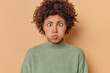 © wayhome.studio - Photo of funny curly haired young woman pouts lips blows cheeks makes hilarious grimace holds breath crosses eyes dressed in casual jumper isolated over beige background tries to amuse someone