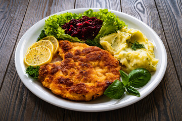 Breaded fried pork chop with potato puree, beetroots, lemon and fresh vegetables on wooden table
