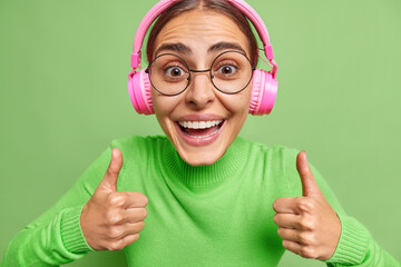 Happy woman enjoys nice audio track does positive gesture with hands keeps thumbs up says I like it wears audio track in pink headphones wears casual clothes round spectaclesisolated on green wall