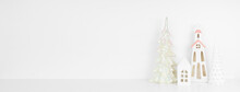 White Christmas Decor On A White Shelf Against A White Wall Banner Background With Copy Space. Ceramic Church, House And Shiny Trees.