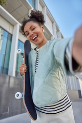 Wall Mural - positive teenage girl with hair buns makes selfie photo of herself holds skateboard enjoys favorite 