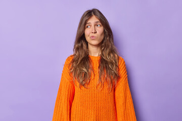 Wall Mural - Photo of thoughtful long haired woman with bored expression hesitates about something purses lips wears knitted orange jumper isolated over purple background. People and face expressions concept