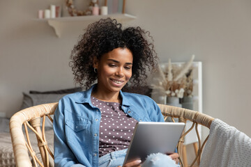 Happy young Black woman reading book, ebook on tablet at home, relaxing in armchair. Smiling girl making video call, chatting online, watching movie, using virtual app on digital tech device