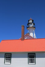 Whitefish Point Lighthouse Behind Red Roof Of Light Keepers Quarter Against Bright Blue Sky