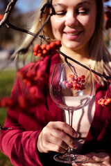 Smiling beautiful blond woman stands with a glass of wine in the sun on the background of rowan branches. Autumn atmosphere and mood