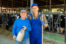Senior Man With Milk Can And Adult Man With Working Tool Standing In Cowhouse And Looking In Camera.