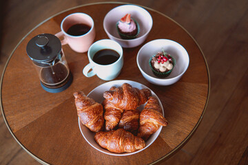 Poster - Closeup shot of cups of coffee with cupcakes and croissant bread in white bowls served in a cafe