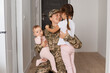 © sementsova321 - Young adult woman soldier wearing camouflage uniform, military female servant meeting his cute lovely girls daughters in the house while coming back home