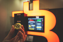 Bitcoin Gold Coin Held In Hand Next To Bitcoin ATM, Orange Cryptocurrency Exchange Machine That Allows To Buy And Sell BTC Crypto. Toned Image With Selective Focus.