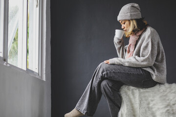 Poster - Lonely girl sitting and drinking a cup of coffee in front of the window on winter season