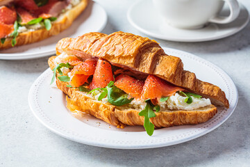 Canvas Print - Croissant sandwich with cream cheese, salmon and arugula on white plate, gray background. Healthy breakfast concept.