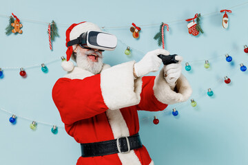 Old cool Santa Claus man in Christmas hat red suit play pc game with joystick console watch in vr headset pc gadget isolated on plain blue background studio Happy New Year 2022 merry ho x-mas concept