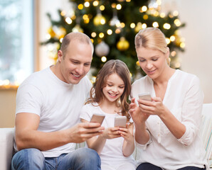 Poster - family, winter holidays and technology concept - happy mother, father and daughter with smartphones at home over christmas tree lights background