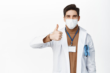 Wall Mural - Covid-19 and pandemic. Smiling doctor in medical face mask, showing thumb up, standing in white robe over studio background