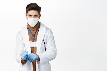 Wall Mural - Covid 19. Doctor in medical face mask, put on gloves for patient examination, wearing clinic uniform, standing over white background