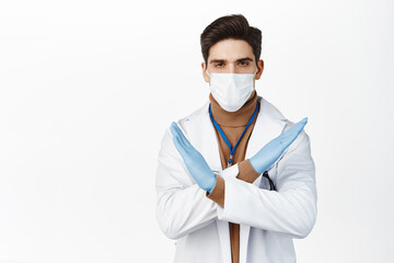 Wall Mural - Young doctor in medical face mask from covid 19, rubber gloves, making cross stop gesture, prohibit action, standing over white background