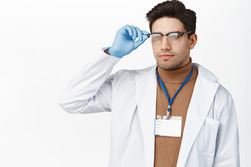 Wall Mural - Healthcare and medical concept. Young male doctor in hospital uniform, wearing glasses and name tag, looking aside with serious face, white background