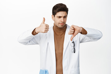 Wall Mural - Indecisive doctor showing thumb up and thumb down, cant decide, making choice, standing in white robe against white background