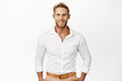 © Cookie Studio - Handsome macho man with blond hair and beard, smiling and looking confident at camera, wearing white shirt, standing against studio background