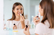 beauty, make up and cosmetics concept - teenage girl applying lipstick and looking to mirror at home bathroom
