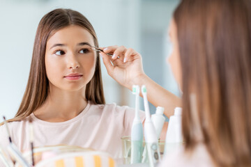 Wall Mural - beauty, make up and cosmetics concept - teenage girl with tweezers tweezing her eyebrow and looking to mirror at home bathroom