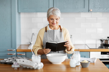 Wall Mural - culinary, baking and people concept - happy smiling woman with cook book cooking food on kitchen at home