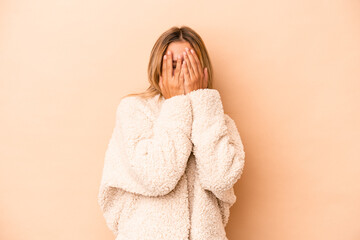 Poster - Young caucasian woman isolated on beige background blink at the camera through fingers, embarrassed covering face.