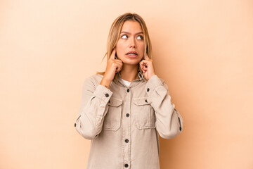 Poster - Young caucasian woman isolated on beige background covering ears with fingers, stressed and desperate by a loudly ambient.