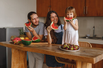 A family in the kitchen cuting and eating the watermelon at home. Happy family of mother, father and lttle daughter eating fruits at home kitchen, have fun wit slices of watermelon