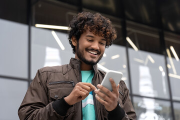 happy young indian man smiling holding mobile phone text social media outdoors