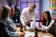 © sofiko14 - Caucasian man in white shirt looking on camera during conference with multiracial partners. Business people sitting at desk office with papers and modern technology.
