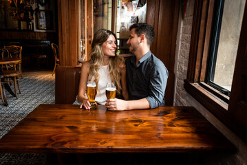 cute and young man and woman sitting down at indoor pub having fun looking at each other smiling and drinking draft beer