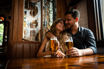 attractive young man and woman couple in love sitting down at pub indoors drinking draft beer and celebrating