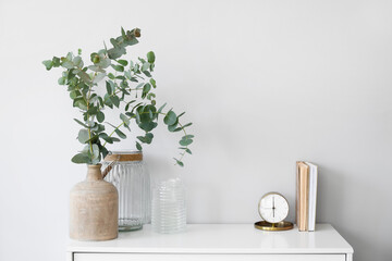 vases with eucalyptus branches, alarm clock and books on shelf near light wall