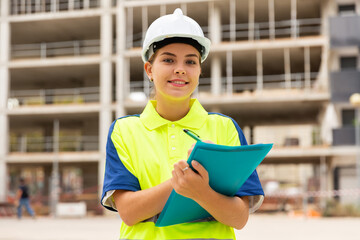 Young attractive girl engineer in hard hat with papers checking work process in construction site outdoors