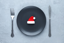 Christmas Table Setting. A Black Plate With A Santa Claus Hat And Silverware On A Gray Background. Top View. Concept: Christmas Dinner.