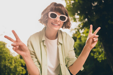 Wall Mural - Photo portrait young girl with bob hair in sunglass showing v-sign gesture on sunny summer street wearing casual clothes