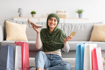 Wall Mural - Arab woman in hijab holding credit card and new shoe, sitting on floor among gift bags, buying footwear on web at home