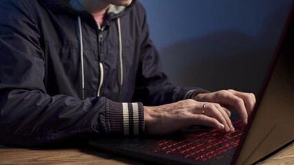 Wall Mural - A hooded hacker is typing on a laptop keyboard in a dark room under a blue light. The concept of cybercrime fraud and identity theft